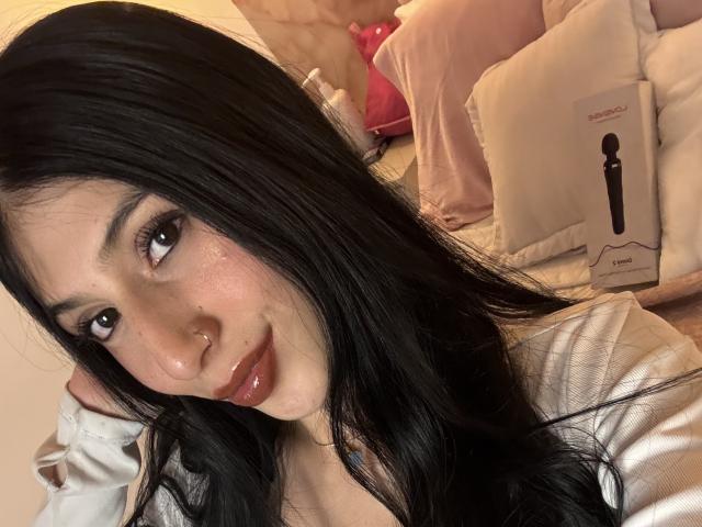 Foto van Sophie Beaumont, close-up, zwart haar, glanzende lippen, roze wangen, draagt een witte blouse, leunt voorover. Hand achter het hoofd, rode lippen, sexy uitdrukking, bed met een roze tas en doos op de achtergrond..