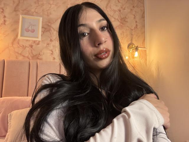 Fotografie von Sophie Beaumont, mit langen, schwarzen Haaren, glänzenden roten Lippen und einem Nasenring, die ein weißes Hemd trägt, Arme in einem rosafarbenen Schlafzimmer mit einer angezündeten Lampe und Blumenmuster-Tapete. Verdammt heiß..