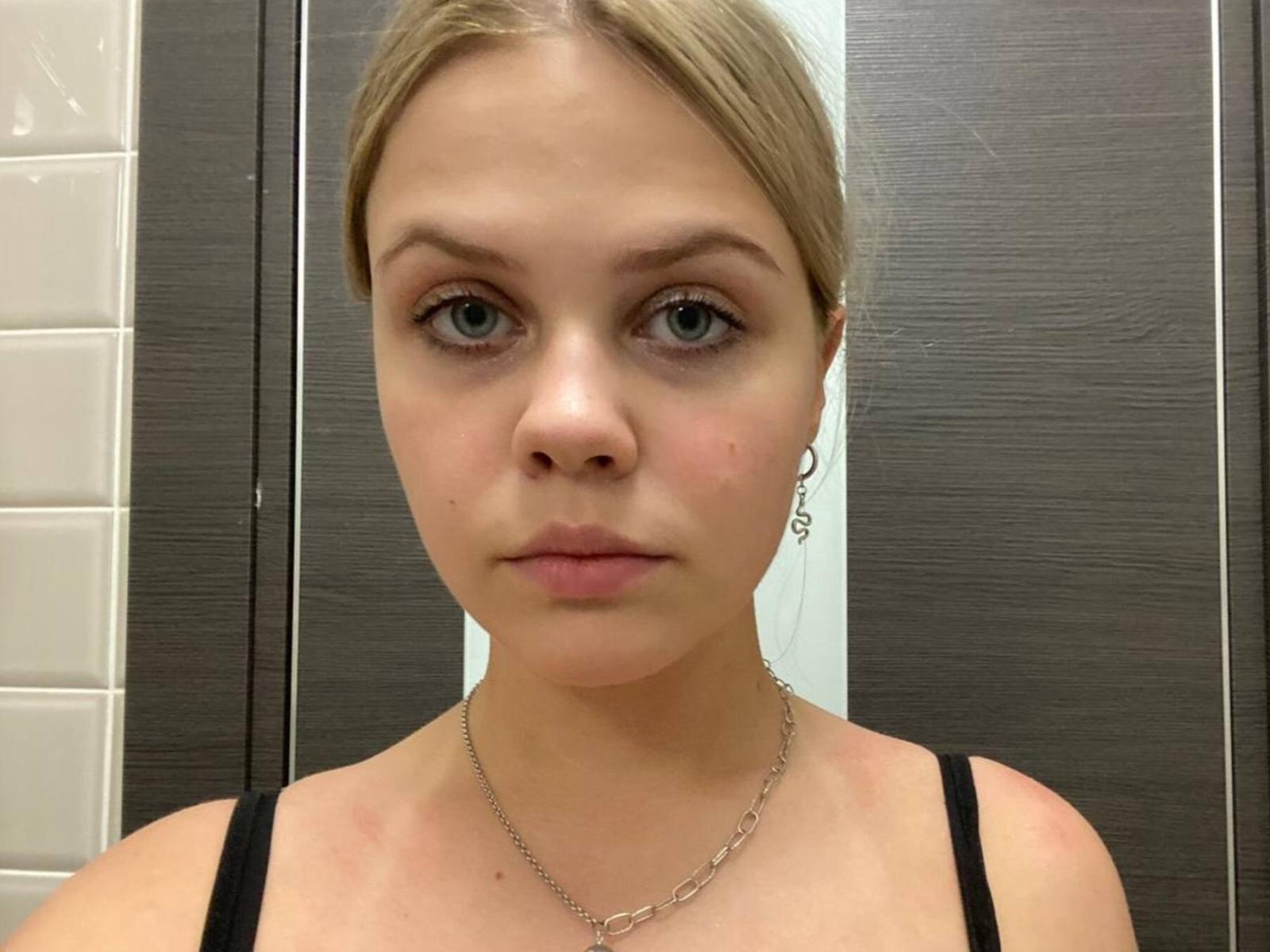AlbaPenninger has blonde hair, blue eyes, and a neutral expression. She's wearing a black top with a silver necklace, and her small tits are slightly visible.