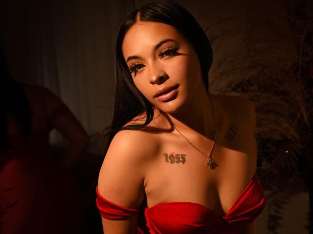 Fotografía de MiaMillerr69, con cabello largo y negro, vistiendo una blusa roja con un hombro descubierto, que muestra un tatuaje que dice "tess" en su pecho. La luz cálida resalta su expresión seductora, con un pezón visible..