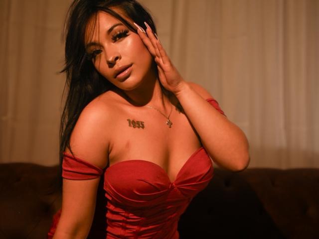 Photograph of MiaMillerr69, dark-haired woman with tan skin, wearing a red off-shoulder dress, hand on face, cleavage accentuated, tattoo on chest, intense gaze, dimly lit, sultry expression.