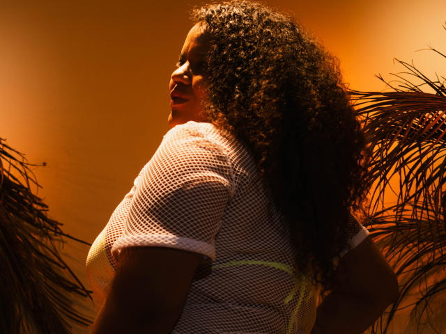 Fotografie von MhiaRey, einer dunkelhäutigen Frau mit lockigem Haar, die ein weißes Netzoberteil trägt und von einem leuchtend orangefarbenen Licht beleuchtet wird. Licht. Palmenblätter im Hintergrund. Ausdrucksstarker, sinnlicher Gesichtsausdruck. Schatten betonen die Kurven..