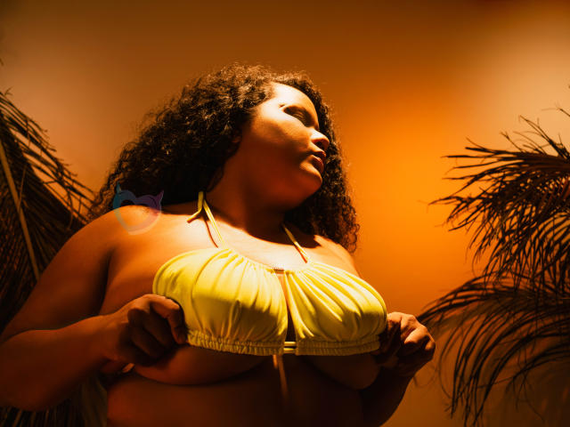 Fotografia de MhiaRey com cabelo cacheado escuro, vestindo um top de alça amarela, segurando seus seios grandes. Brilho alaranjado, folhas de palmeira ao fundo. Foco na sua pose sexy e confiante..