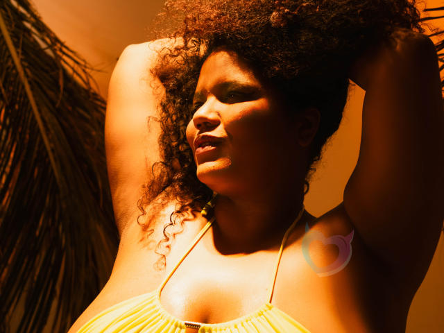 Close-up foto van MhiaRey met donkerbruine huid en krullend haar, armen omhoog, gekleed in een geel topje met halter, verlicht door intens oranje licht, dat scherpe schaduwen creëert en een sensuele sfeer..