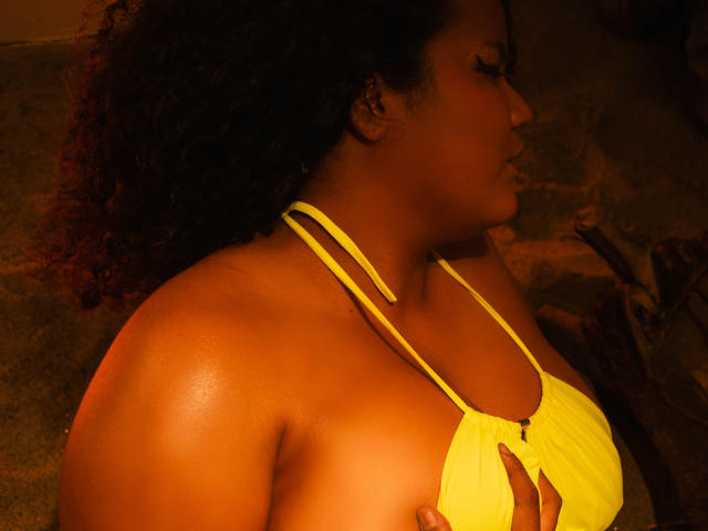 Photograph of MhiaRey with dark skin, curly hair, and a yellow bikini top, holding her large breasts. Bright orange lighting highlights her sweaty, glistening skin. Profile view, side light.