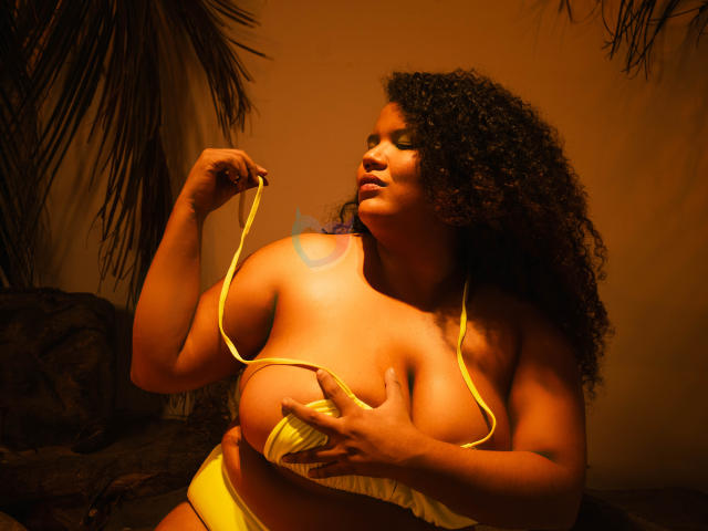 Photograph of MhiaRey with dark curly hair, wearing a yellow bikini, holding one strap, eyes closed, light glowing on her skin, palm fronds in background, intense orange lighting. Fucking hot.
