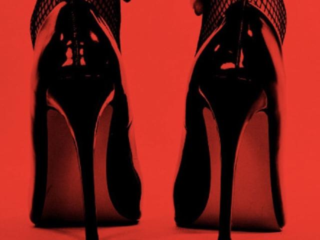 Close-up foto van SofiBrandford's zwarte stiletto pumps met puntige neus en hoge hakken, op een achtergrond van felrood. Achtergrond. Zwarte maasbroek, gedeeltelijk zichtbaar. Intense, erotische sfeer..