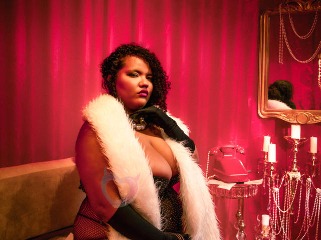 MhiaRey, avec ses cheveux noirs et bouclés, portant une robe noire à décolleté plongeant et une écharpe en fourrure blanche, assise dans une pièce éclairée en rouge. Bougies rouges et miroir en arrière-plan. Pose sexy et confiante..