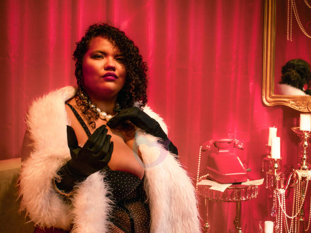 MhiaRey with curly black hair, wearing a black sparkly dress, white fur coat, and black gloves, stands in a red-lit room, holding the coat with a seductive gaze. Red candles nearby.