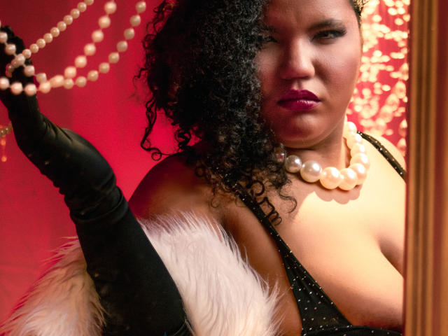 Foto em close-up de MhiaRey, uma mulher de pele escura com cabelo preto encaracolado, usando luvas pretas, um colar de pérolas e preto blusa com bolinhas e detalhes de pele branca, segurando um colar de pérolas, fundo vermelho. Muito atraente..