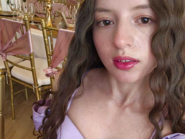 Close-up of VioletaNova with long wavy brown hair, pink lipstick, and a white top. She's in a room with gold chairs and pink sashes. Fucking hot, sexy, and slightly flushed.