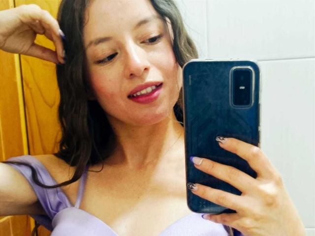 Primo piano di VioletaNova, una donna dalla pelle chiara con i capelli castani bagnati, che si scatta un selfie con un telefono nero. Indossa una maglia bianca con spalline viola, rossetto rosso e ha un'espressione seducente..
