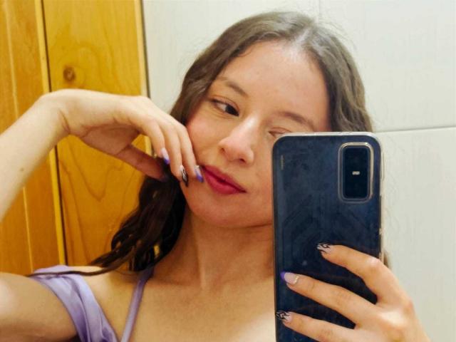 Autofoto de VioletaNova, sosteniendo un teléfono inteligente negro en su mano derecha, con su mano izquierda tocando sus labios rojos. Ella tiene el pelo largo y castaño, piel clara y lleva una blusa color lavanda..