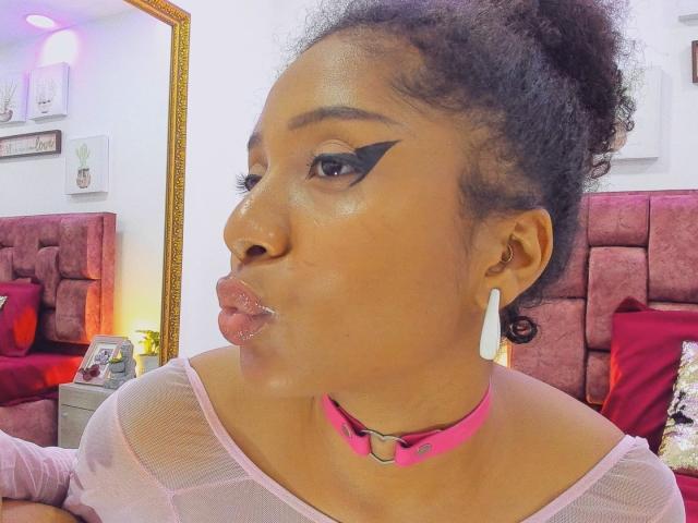 Close-up photo of MeganWilliams with dark skin, thick black hair in a bun, wearing bold black eyeliner, pink choker, white mesh top, puffy lips, and white earrings. Background has red headboard.