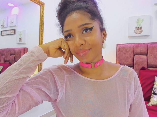 Megan Williams, con la pelle di un marrone medio, i capelli neri ricci raccolti in uno chignon, indossa una maglia rosa trasparente e una collana choker rosa acceso. con la testa appoggiata sulla mano, guarda direttamente la fotocamera con un sorriso appena accennato..