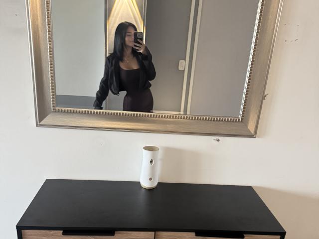 Selfie no espelho de MajoJones, com cabelo preto longo, vestido preto, seios grandes, segurando o celular, reflexo em um espelho ornamentado. mesa preta com caneca branca, paredes cinzas, tensão sexual, pose casual.