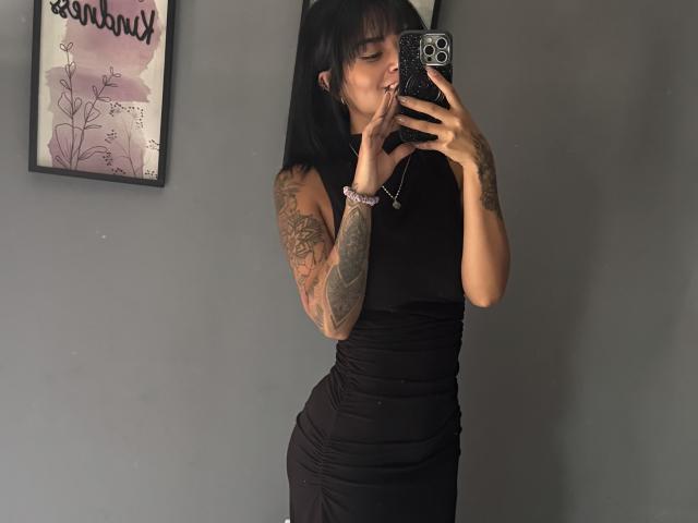 MajoJones macht ein Selfie vor einem Spiegel und trägt ein eng anliegendes schwarzes Kleid, wobei ihre tätowierten Arme zu sehen sind. Sie hält ein Telefon mit einer weißen, gemusterten Hülle in der Hand und betrachtet sich dabei mit leicht geöffnetem Mund..