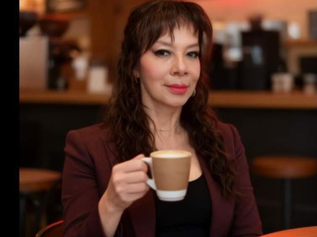 Photograph of SamaraBellet, brunette, wavy hair, red lipstick, black blazer, holding a cappuccino, slight smile, bar stool blurred background, warm lighting, sexy, confident, casual cafe setting, slight blur on edges.