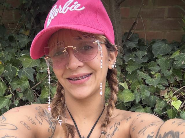 Nahaufnahme von CassieInk mit Tattoos an Schultern und Brust, die eine rosa "Nike"-Kappe und klare Aviator-Sonnenbrillen trägt. und Nasenring. CassieInk hat ein verschmitztes Lächeln und ein sichtbares Tattoo am Schlüsselbein. Im Hintergrund ist grünes Laub zu sehen..