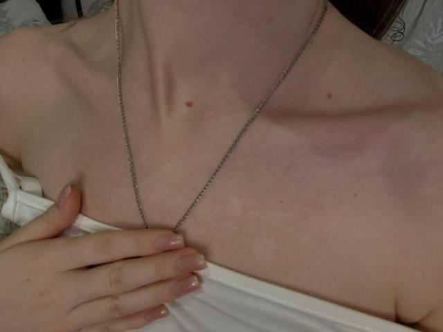Close-up of HanHeffren's chest, showing pale skin with small moles, wearing a thin silver necklace and a white top pulled down slightly. HanHeffren's hand with pink nails touches collarbone.