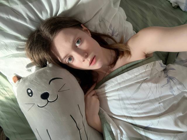 HanHeffren, acostada en la cama, con los ojos medio abiertos, mirando hacia arriba. Cabello castaño, piel pálida, ojos verdes. Sosteniendo un cojín blanco con la cara de un gato sonriente. Debajo de una manta verde y blanca, mostrando el hombro. Luz del sol en el rostro.