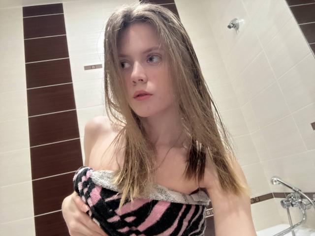 Fotografía de HanHeffren con el cabello castaño, mojado y de largo medio, sosteniendo una toalla a rayas negras y rosas sobre su pecho. Está en un baño luminoso con azulejos blancos y marrones. Un aspecto húmedo, con el pene apenas cubierto..