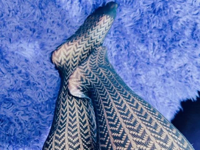 Primo piano delle gambe di Cincia69, che indossa calze a motivi a zig-zag bianche e nere, sdraiate su un tappeto blu e peloso. La sua gamba destra è incrociata sopra la sinistra, creando una posa sensuale e provocante..