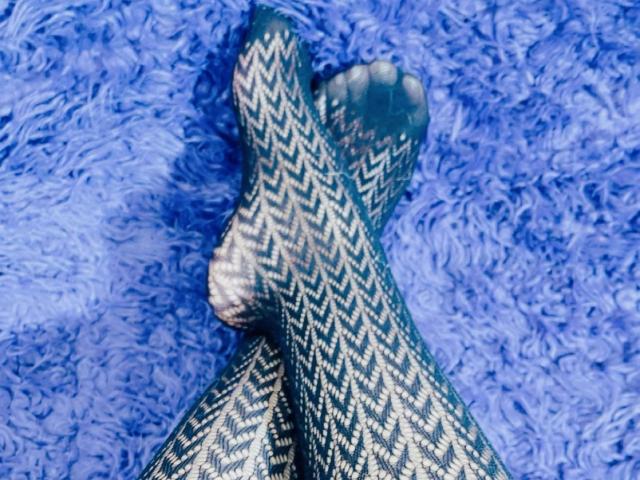Nahaufnahme von Cincia69s Beinen, die auf einem blauen, flauschigen Teppich gekreuzt sind. Sie trägt schwarz-weiße, zickzackmusterte Strumpfhosen. Ihre Zehen sind sichtbar, und die Textur des Teppichs ist weich. Verdammt sexy..