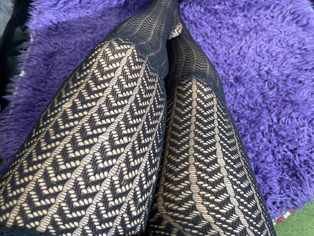 Gros plan des jambes de Cincia69, portant des bas de pêche noirs à motifs, croisées sur une couverture violette et duveteuse. Le motif en filet crée un effet de treillis, soulignant la texture et la tension..