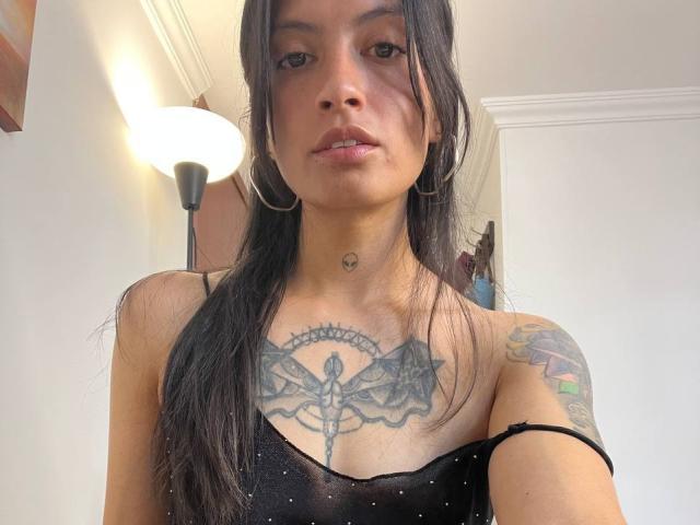 Primo piano di NiaWhitee, con lunghi capelli neri, che indossa una canottiera nera a spalline sottili, che rivela un grande tatuaggio sul petto. design con elementi alari.
tatuaggio visibile sulla spalla sinistra, espressione seria.