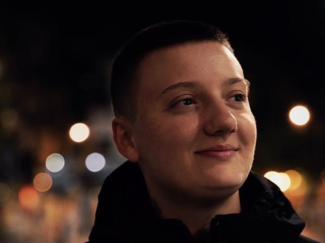 Photographie de JamesChet, avec une courte coupe de cheveux, une peau claire et un léger sourire. Il porte une veste sombre, avec des lumières de ville floues en arrière-plan. Légère tension sexuelle.