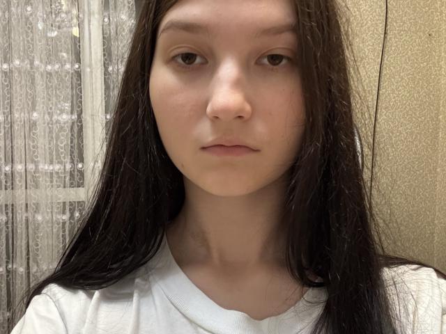 Close-up of DrewHugo with long black hair, pale skin, and neutral expression. Wearing a white shirt, standing in front of a textured wall and patterned window. Slight sexual tension.