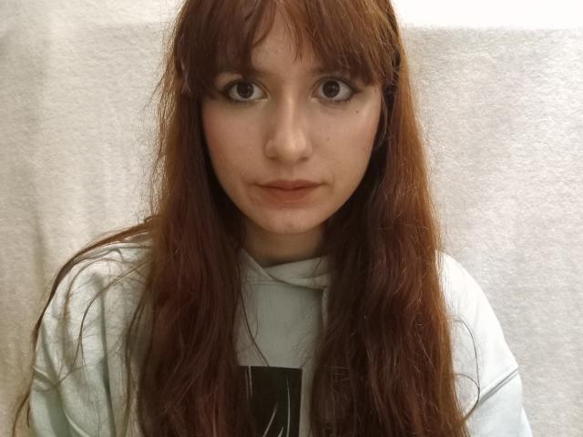 Close-up van GregoriaKubat, een jonge vrouw met lang bruin haar, grote bruine ogen en een serieuze uitdrukking. Ze draagt een witte hoodie, met een helder licht op de achtergrond. Extreem intense blik.