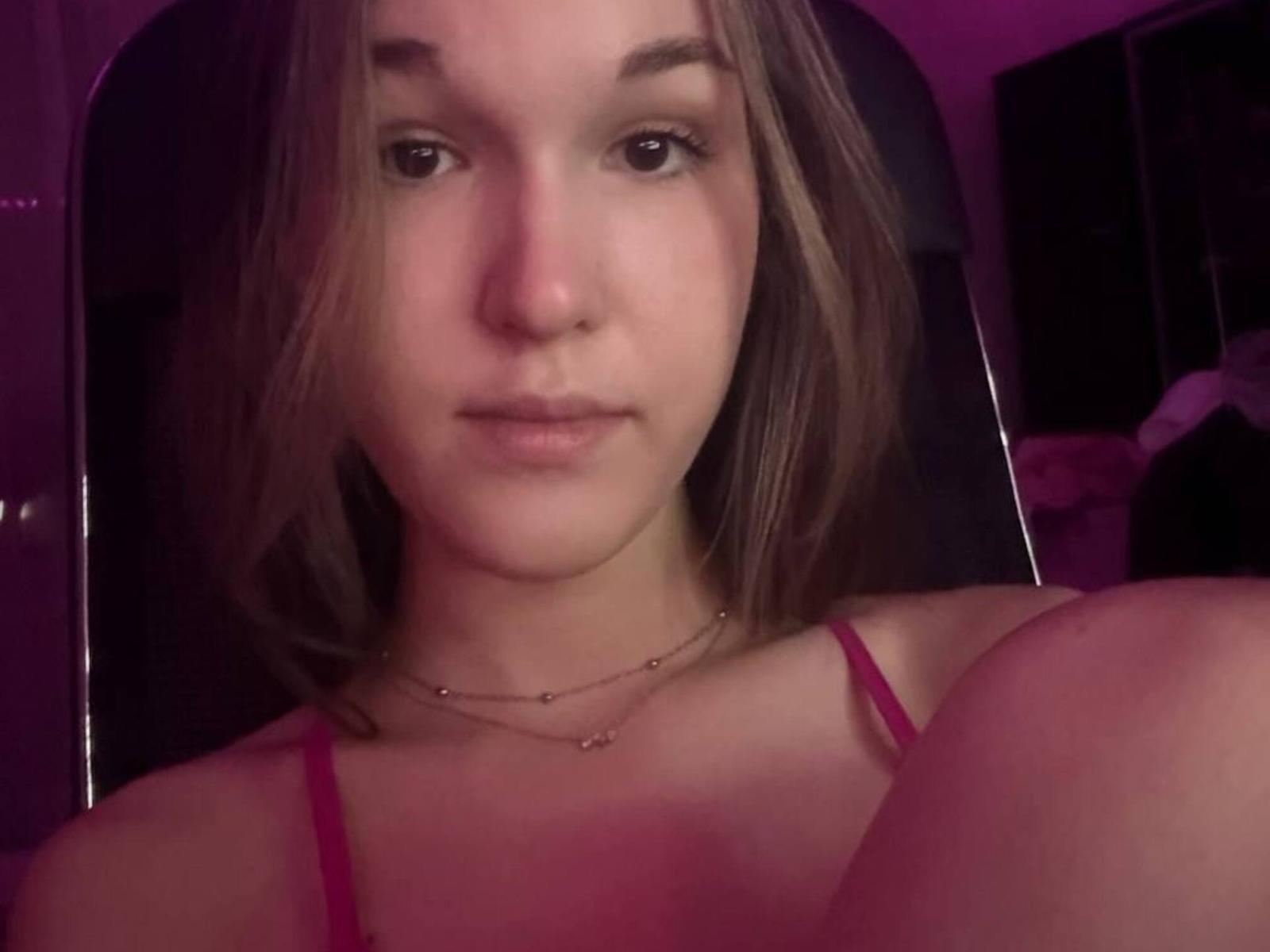 Photo en gros plan de PhungEmmert, une jeune femme à la peau claire et aux cheveux bruns, portant un haut rouge à bretelles fines, qui regarde directement face à la caméra, avec une expression neutre. Éclairage rosé.