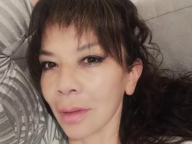Close-up photo of SamaraBellet, lying down with dark, wavy hair and bangs. She has light skin, dark eyes, and pink lips. Gray pillow with pattern in background. Fucking sexy expression.