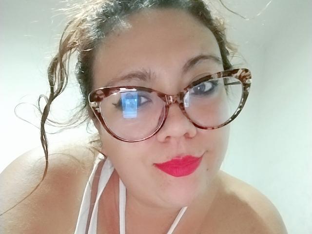 Fotografía de primer plano de LexyDom, una mujer robusta con cabello castaño, que lleva gafas de montura carey y un lápiz labial rojo brillante. mirando directamente a la cámara. Ella lleva un top blanco con tirantes, y tiene una expresión ligeramente seductora..