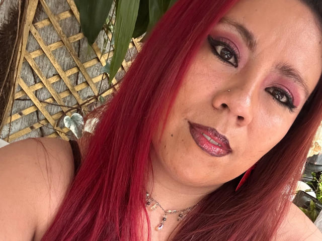 Fotografía de primer plano de LexyDom con cabello largo y rojo, sombra de ojos rosa intensa y labios rosados brillantes. Ella lleva un collar, y su rostro muestra una sonrisa sutil. Se ve un enrejado de madera y vegetación en el fondo..