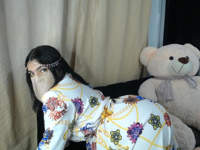 AniisaAli with black hair, wearing a white floral kimono, fishnet face mask, and headpiece, leans forward, showing ample ass. Large white teddy bear on the right. Sexual tension, slutty vibe.