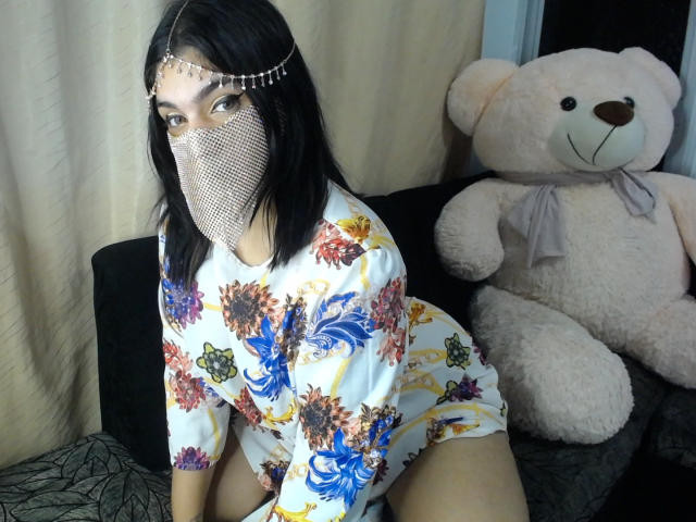 Aniisa Ali, con i capelli neri, indossa un abito bianco con fiori, una maschera a rete e una catena per i capelli, seduta su una sedia nera accanto a un grande orsacchiotto bianco con un fiocco viola.