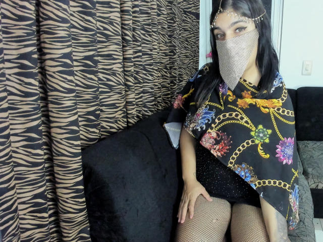 AniisaAli sits on a black couch, wearing a floral black shawl with gold chains, fishnet stockings, and a white mesh face mask. Zebra-patterned curtains are in the background. Fucking sexy.