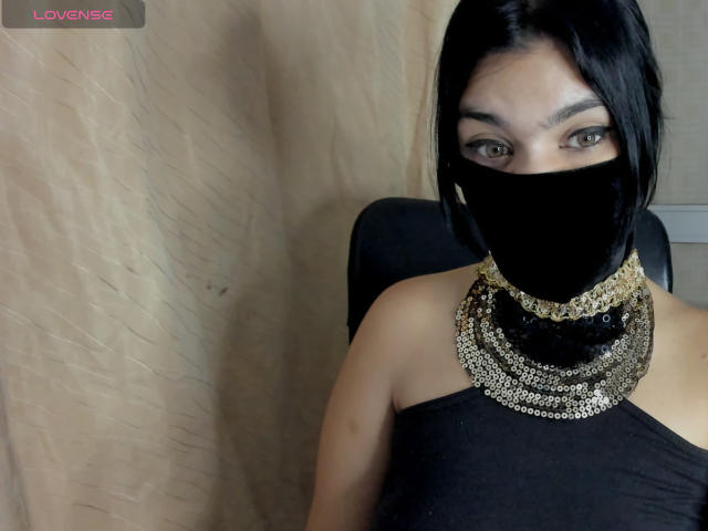 Fotografia de Aniisa Ali, uma mulher asiática com cabelo preto, vestindo uma blusa preta sem alças, um colar dourado e uma máscara facial preta. Ela olha diretamente para a câmera, transmitindo uma intensa tensão sexual. "AMOR" no canto superior esquerdo.