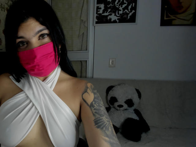 AniisaAli, con cabello oscuro, máscara roja, top blanco con diseño cruzado que deja ver el escote, brazo tatuado, sentada en un sofá. Peluche de panda en blanco y negro. Habitación con poca luz, arte en las paredes en blanco y negro. Increíblemente sexy..
