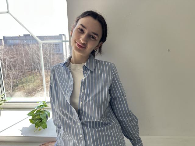JessicaBreeze está apoyada contra una pared blanca, vestida con una camisa de rayas azules y blancas sobre una camiseta blanca. La luz del sol entra por una ventana detrás de ella, resaltando su sonrisa. Su cabello oscuro está recogido. Una planta en maceta está sobre el alféizar de la ventana..