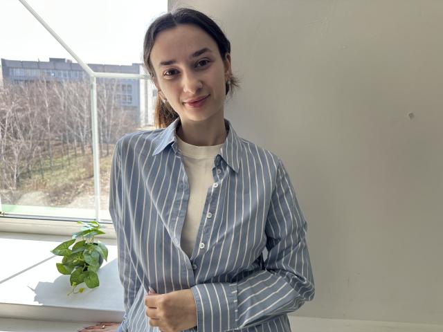 JessicaBreeze, parada em frente a uma janela, vestindo uma camisa de botões listrada em azul e branco por cima de uma camiseta branca, sorrindo levemente.
luz vindo da janela, planta no parapeito. Casual, ligeiramente sensual..