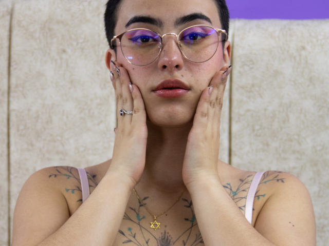 Foto em close-up de CatsitoS, uma pessoa tatuada com cabelo curto e escuro, usando óculos redondos com lentes azuis e uma regata branca. parte superior do corpo, colar de ouro, mãos no rosto, olhar intenso, tatuagens visíveis no peito..