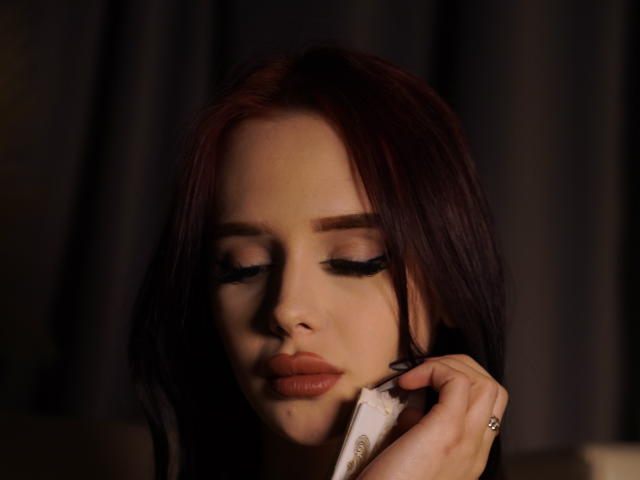 AleneNghiem met rood haar, gesloten ogen en volle lippen, die een telefoon op haar oor houdt, zwarte eyeliner draagt en een ring om heeft, in een omgeving met weinig licht..