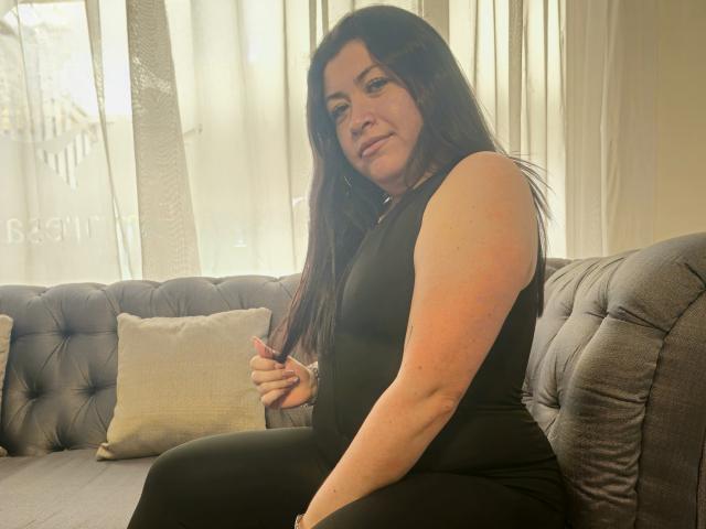 Ayleen Miller, avec ses longs cheveux noirs, est assise sur un canapé gris capitonné, vêtue d'une robe noire moulante sans manches, et tient cigarette, rayons de soleil filtrant à travers des rideaux blancs, tension sexuelle, pose détendue.