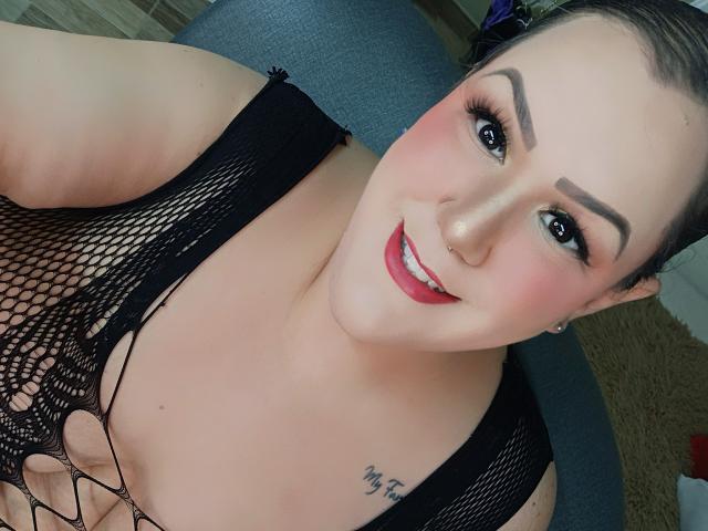 Close-up photo of GiiannaMorrys, smiling with red lipstick, black fishnet top, revealing cleavage, visible tattoo on right shoulder. Dark eyebrows, blue eyes, nose piercing. Seductive, confident expression.