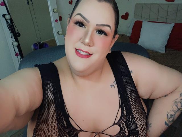 Close-up photo of GiiannaMorrys, a plus-sized woman with fair skin, dark hair in a bun, red lipstick, and black fishnet lingerie. She's smiling, showing a tattoo on her arm, with hearts in the background.