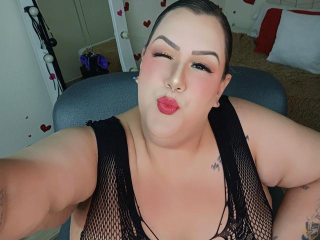 GiiannaMorrys, overweight, pale-skinned, black fishnet top, dark hair slicked back, septum piercing, red lipstick, winking, slightly open mouth, arm tattoos, grey chair, bed with red and white pillows in background. Fucking sexy.